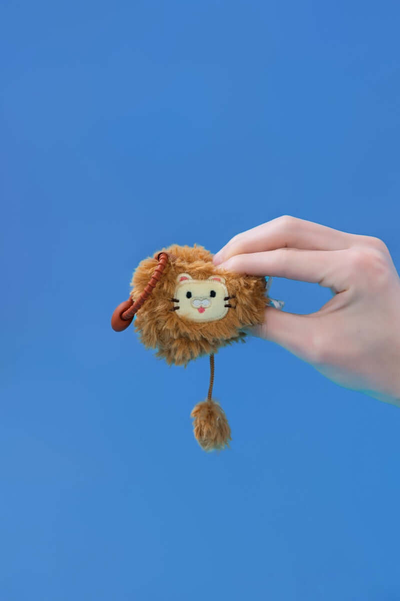 Mini lion plush toy being held against a blue background, featuring a cute design and clip-on strap for easy attachment.