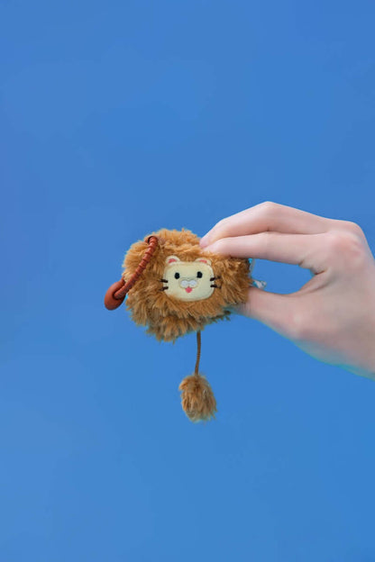Mini lion plush toy being held against a blue background, featuring a cute design and clip-on strap for easy attachment.