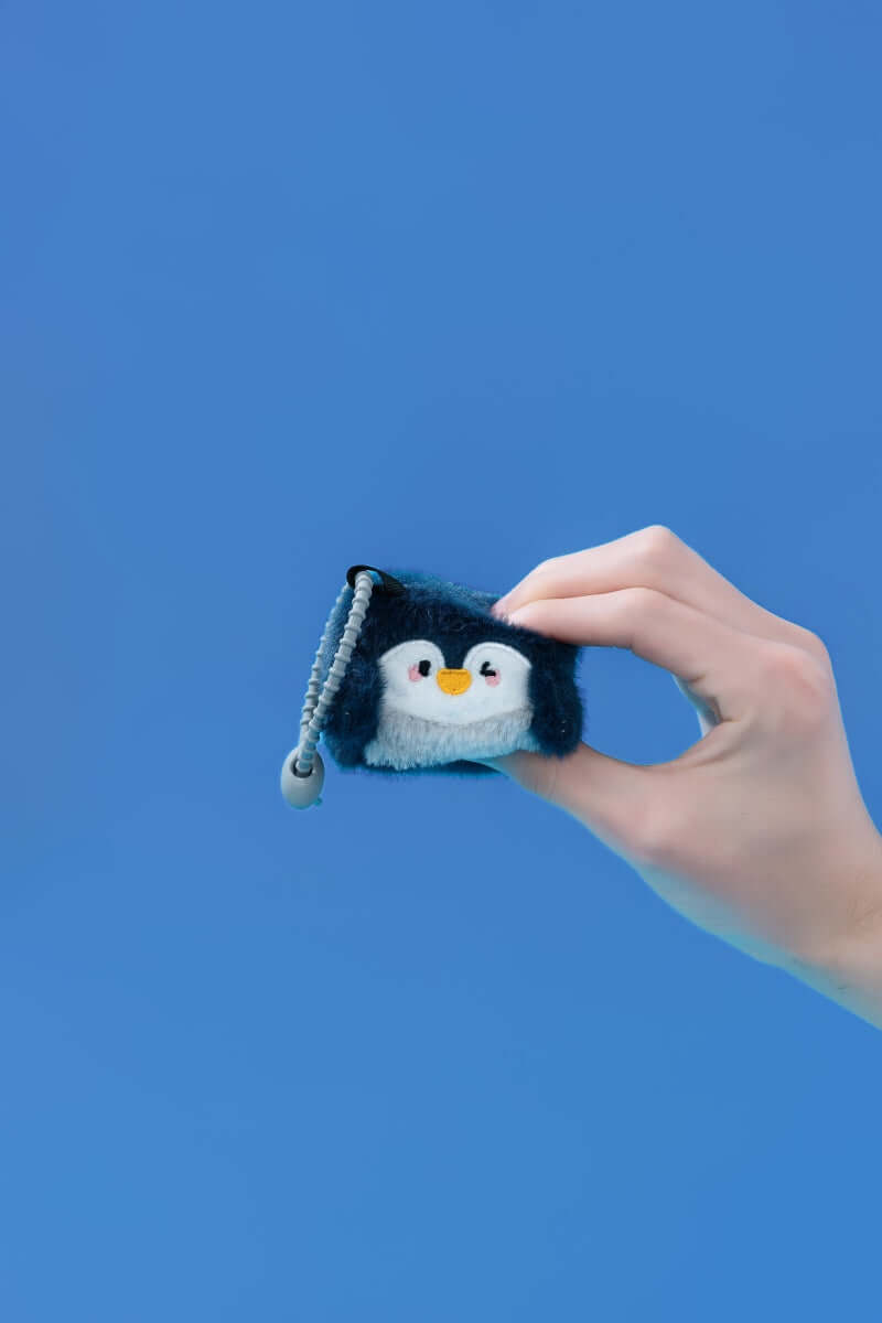 Hand holding a mini penguin plush toy with a clip-on strap against a blue background.