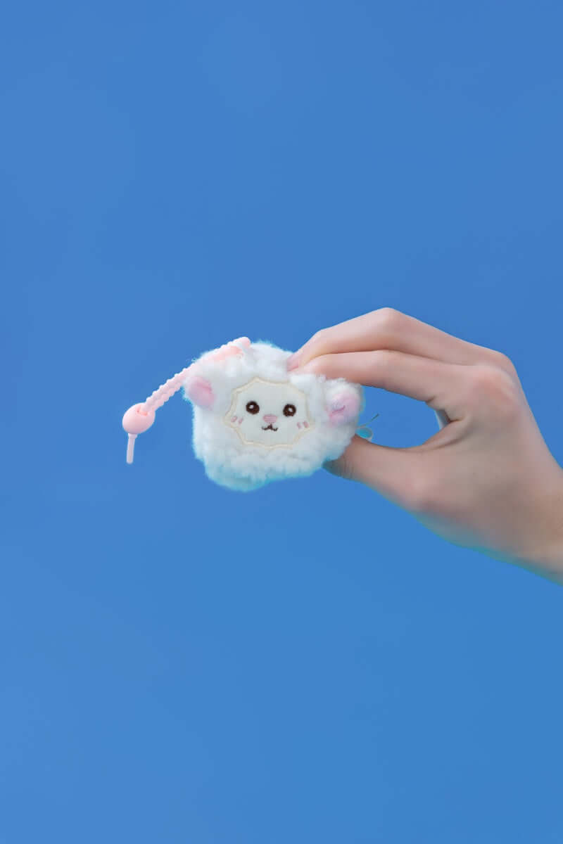 Mini squishy sheep plush toy held in hand against a blue background, showcasing its cute design and clip-on feature.